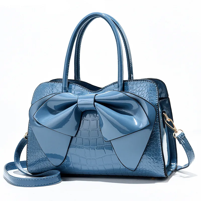 Denim croc-embossed handbag with a glossy front bow, two handles, and a long strap.